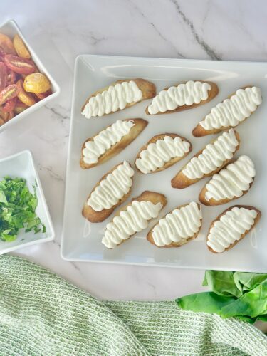 A plate of baked bread slices topped with white cream, arranged neatly. Surrounding the plate are small bowls of chopped cilantro and colorful roasted vegetables, with a green cloth partially shown.