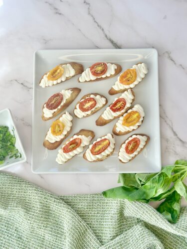 A plate of sliced bread topped with creamy white frosting and colorful tomato slices, arranged artistically on a marble surface, alongside fresh herbs and a green dish towel.