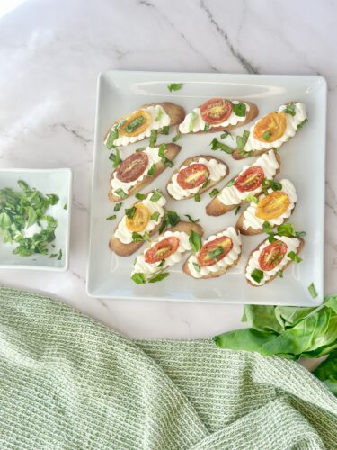 A platter of toasted bruschetta topped with cream cheese, colorful cherry tomatoes, and fresh herbs, accompanied by a small dish of chopped green onions.