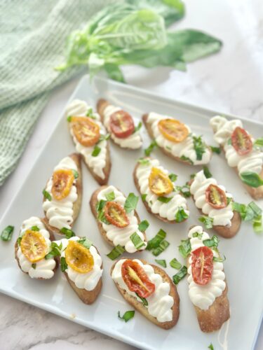 A platter of appetizer bites featuring small toasted bread topped with creamy white frosting, garnished with sliced cherry tomatoes and fresh green herbs on a light background.