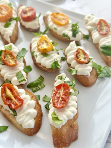 A plate of bruschetta topped with creamy white cheese, fresh basil, and halved cherry tomatoes, arranged neatly.