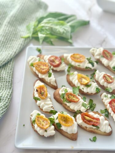Platter of white bread slices topped with cream cheese, fresh herbs, and colorful roasted cherry tomatoes, with a green cloth and basil leaves in the background.