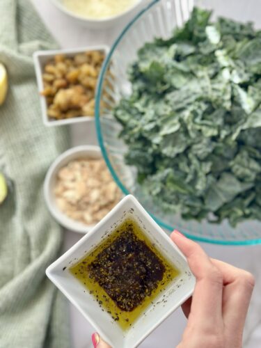 A hand holding a small dish of olive oil dressing, with a bowl of freshly chopped kale in the background and several small bowls containing nuts and cheese.