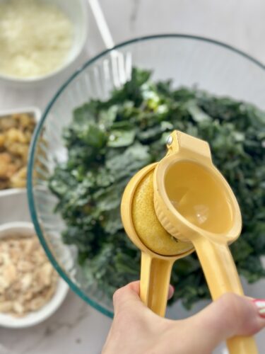 A hand holding a yellow lemon squeezer above a bowl of chopped kale, with small bowls of ingredients in the background.