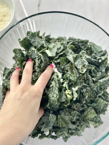 A hand reaching into a glass bowl filled with fresh, leafy kale.