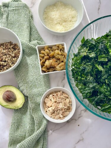 A collection of ingredients for a salad including chopped kale in a glass bowl, quinoa, raisins in a small dish, sliced almonds in another dish, shredded cheese in a white bowl, and half an avocado, set on a marble surface with a green kitchen towel.