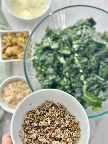 A hand holding a small bowl of ground nuts and seeds, with a large bowl of chopped kale in the background, alongside small bowls of grated cheese, mixed nuts, and shredded chicken.