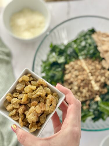 A hand holding a small bowl of golden raisins in front of a glass bowl filled with chopped greens and other ingredients.