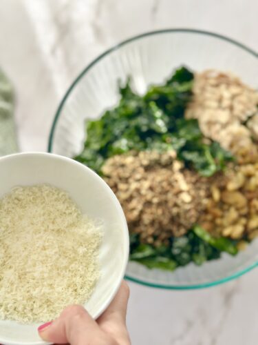 A close-up of a hand holding a bowl of grated cheese over a large glass bowl filled with mixed greens, nuts, and grains.