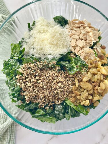 A glass bowl filled with a mixed salad of kale, topped with grated cheese, sliced almonds, ground nuts, and mixed dried fruits.