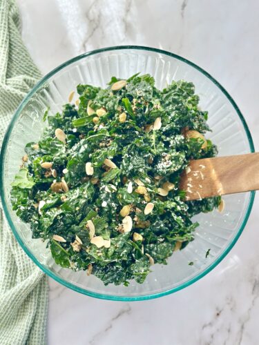 A bright green kale salad with sliced almonds and a wooden spoon in a clear glass bowl on a marble countertop.