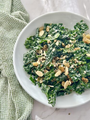 A plate of kale salad topped with sliced almonds, raisins, and a light dressing, placed on a marble surface next to a green kitchen towel.