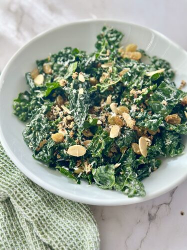 A bowl of kale salad topped with almond slices, dried fruit, and a creamy dressing, served on a marble countertop with a green cloth beside it.