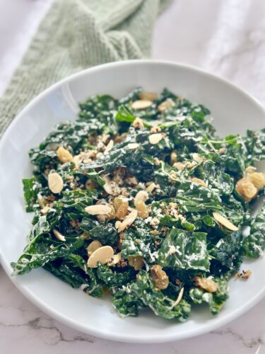 A bowl of kale salad topped with sliced almonds, raisins, and a sprinkle of grated cheese, with a light green textured cloth in the background.