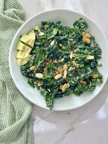A vibrant green kale salad topped with sliced avocado, almonds, and raisins, served in a white bowl on a marble surface.
