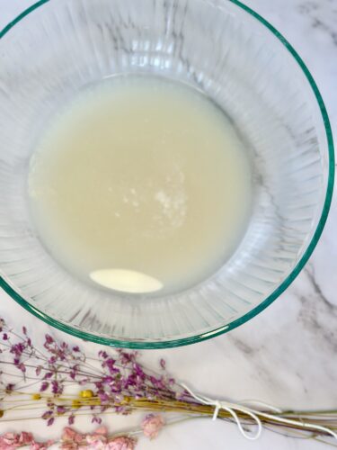 A clear glass bowl filled with a light, translucent liquid, placed on a marble surface next to a cluster of dried flowers.