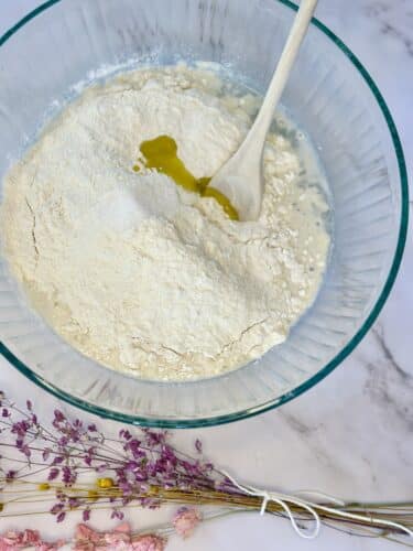 A clear bowl filled with flour and a drizzle of oil, accompanied by a wooden spoon, with a backdrop of dried flowers.
