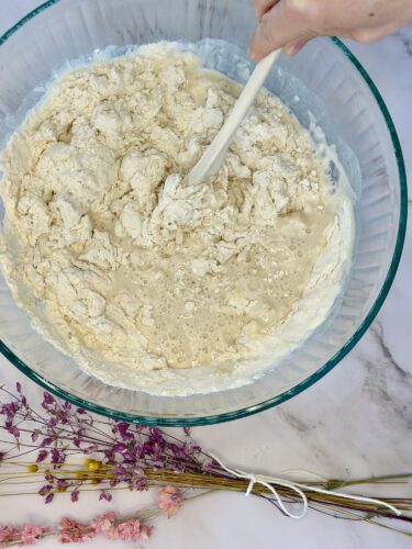 A glass bowl filled with a mixture of flour and wet ingredients, being stirred with a white spatula. In the foreground, there are dried flowers arranged beside the bowl.