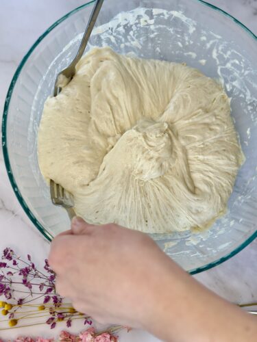 A hand mixing dough in a glass bowl, with soft, fluffy texture, surrounded by dried flowers.