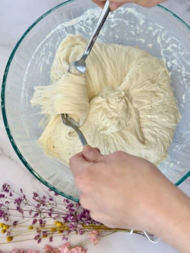 A close-up of hands mixing dough with two forks in a clear mixing bowl, surrounded by dried flowers.