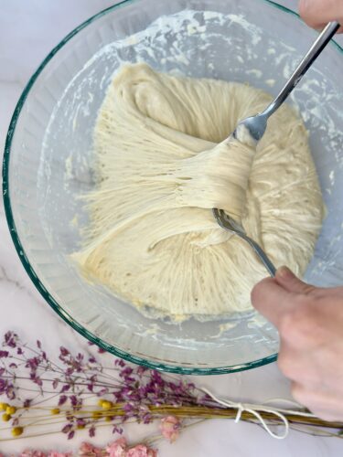 A person mixing a thick, elastic dough in a glass bowl with two forks, surrounded by dried flowers.