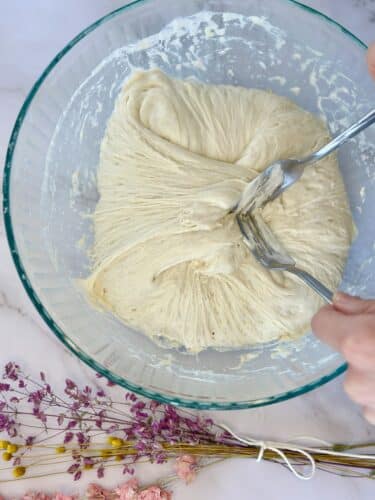 A person mixing dough in a glass bowl with two forks, surrounded by dried flowers.