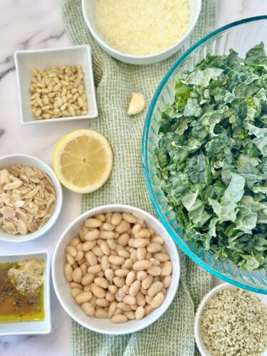A collection of ingredients for a salad, including chopped greens in a large bowl, white beans, sliced almonds, sunflower seeds, lemon, and a small bowl of seasoning, arranged on a green cloth.