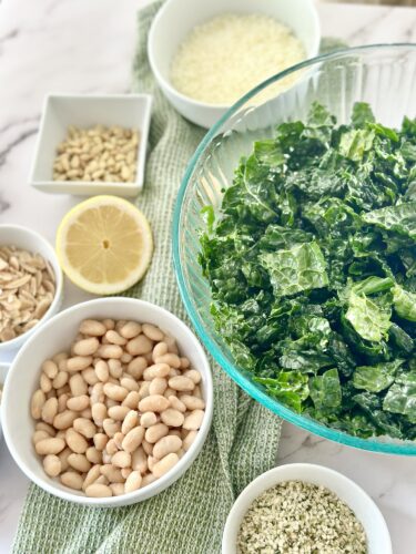A clear glass bowl filled with chopped kale, surrounded by bowls of white beans, sunflower seeds, pine nuts, shredded cheese, a lemon slice, and a small bowl of herbs on a patterned green towel.