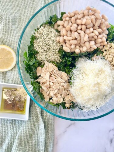 A glass bowl filled with chopped kale, white beans, sunflower seeds, sliced almonds, grated Parmesan cheese, and hemp seeds, with a lemon slice and a small dish of olive oil and garlic in the background.