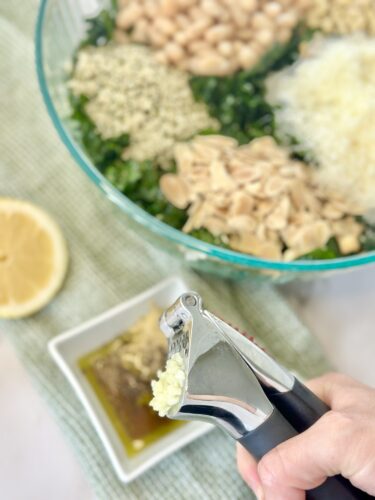 A hand using a garlic press to crush garlic over a bowl filled with various ingredients, including chopped kale, white beans, and sliced almonds, with a lemon and a small dish of olive oil in the background.
