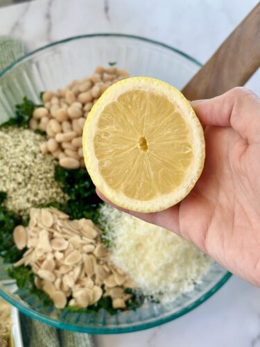 A hand holding a halved lemon above a glass bowl filled with various ingredients including kale, white beans, almond slices, and grated cheese.