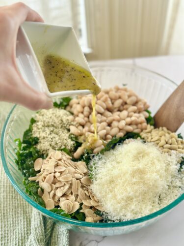 A hand pouring dressing over a salad bowl filled with kale, white beans, cheese, seeds, and almonds.