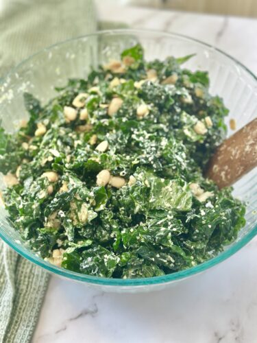 A bowl of kale salad mixed with creamy dressing and topped with pine nuts, with a wooden spoon for serving.