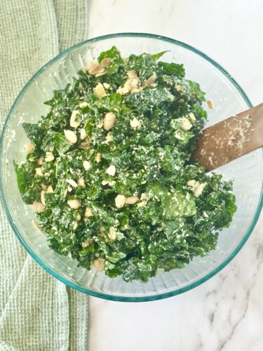 A bowl of kale salad with chopped nuts and a creamy dressing, accompanied by a wooden spoon.