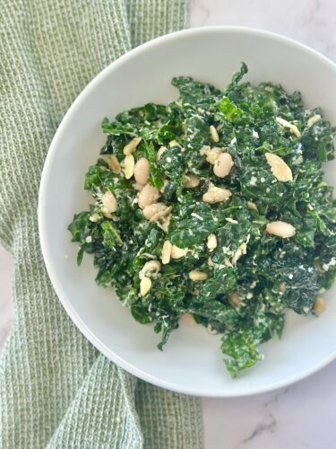 A bowl of kale salad topped with white beans and a creamy dressing, placed on a light green textured cloth.