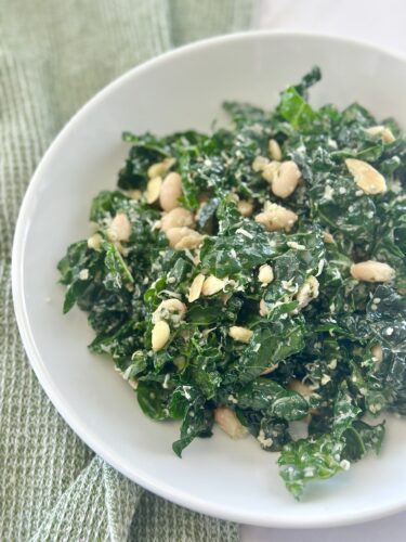 A bowl of kale salad topped with white beans and slivered almonds, resting on a light green cloth.