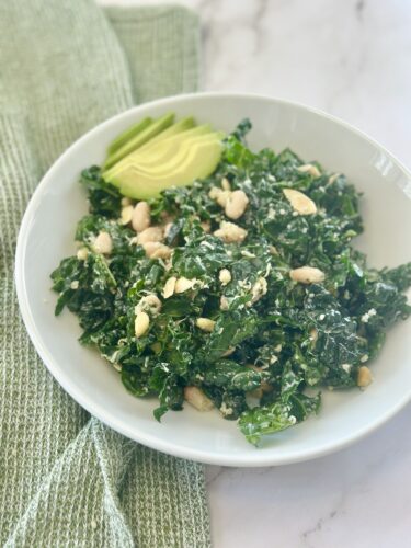 A bowl of kale salad topped with avocado slices and chopped nuts, sitting on a marble countertop with a green napkin beside it.