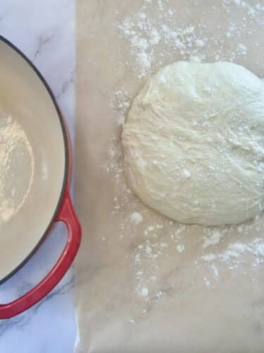 A ball of dough on a floured surface next to a red enameled cast iron pot.