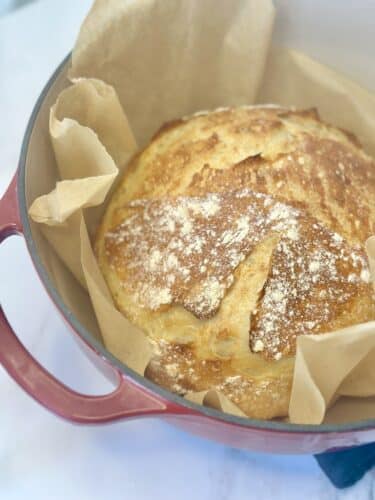 Freshly baked round loaf of bread in a red cast iron pot lined with parchment paper.