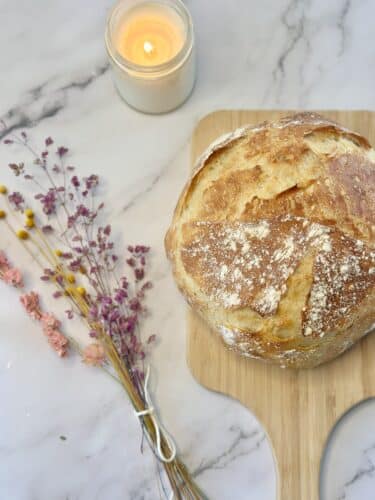 A freshly baked round loaf of bread on a wooden cutting board, surrounded by dried flowers and a lit candle.