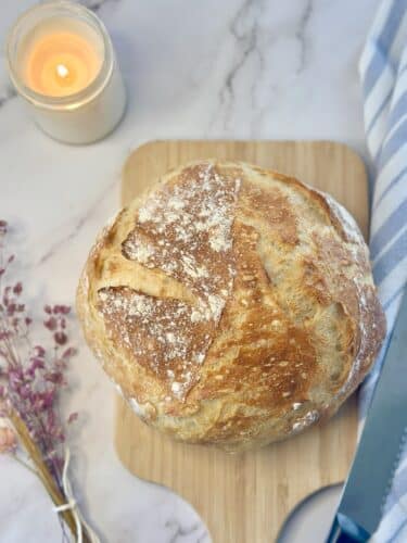 A freshly baked round loaf of bread on a wooden cutting board, with a lit candle and decorative flowers in the background.