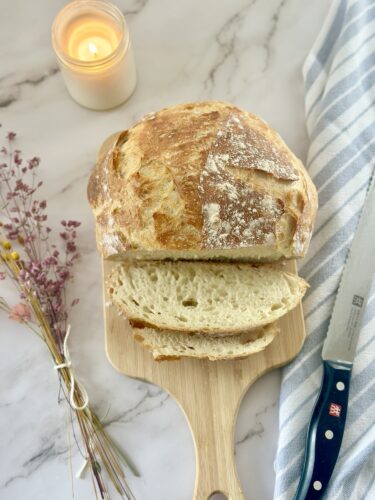 A freshly baked loaf of bread on a wooden cutting board, partially sliced. A lit candle and a knife with a blue handle are beside it, along with some dried flowers.