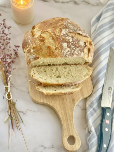 A freshly sliced loaf of crusty bread on a wooden cutting board, accompanied by a candle and a blue-striped dish towel.