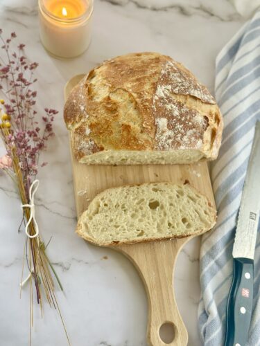 Freshly baked sourdough bread on a wooden cutting board, with a slice cut out, next to a candle and decorative flowers.
