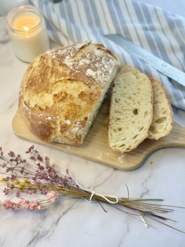 A freshly baked loaf of bread with two slices cut, displayed on a wooden board. A lit candle and a striped cloth are in the background, along with a bundle of dried flowers.