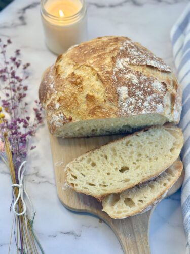 A freshly baked crusty loaf of bread with two slices cut, resting on a wooden board. Nearby, there is a lit candle and a sprig of dried flowers.
