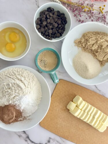 An overview of baking ingredients arranged on a marble countertop, including eggs, chocolate chips, brown sugar, white sugar, flour, and butter slices, along with a cup of coffee.