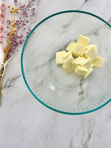 A clear glass bowl containing several cubes of butter placed on a marble surface, accompanied by a bouquet of dried flowers on the side.
