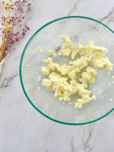 A glass bowl containing fluffy butter with a smooth texture, placed on a marble surface. Dried flowers are visible beside the bowl.