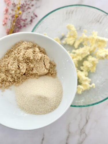 A bowl containing brown sugar and granulated sugar next to a glass bowl of creamed butter mixture, with some dried flowers in the background.